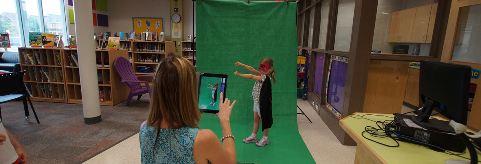 Female adult taking a picture of a female student in front of the green screen